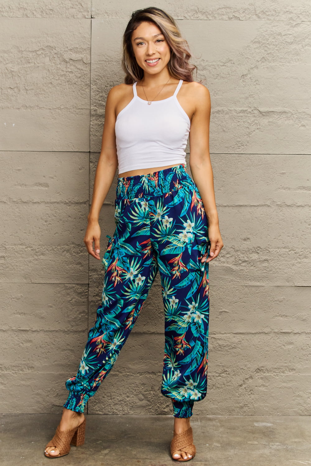 Woman wearing tropical print pants and a white top against a stone wall.