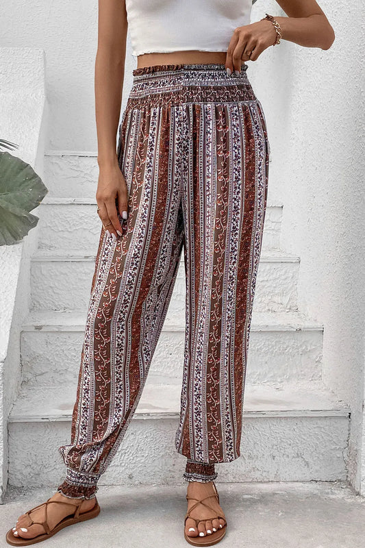 A person wearing loose, high-waisted patterned pants with brown, white, and rust vertical stripes, paired with a white cropped top and brown strappy sandals, standing on white stairs against a textured wall.